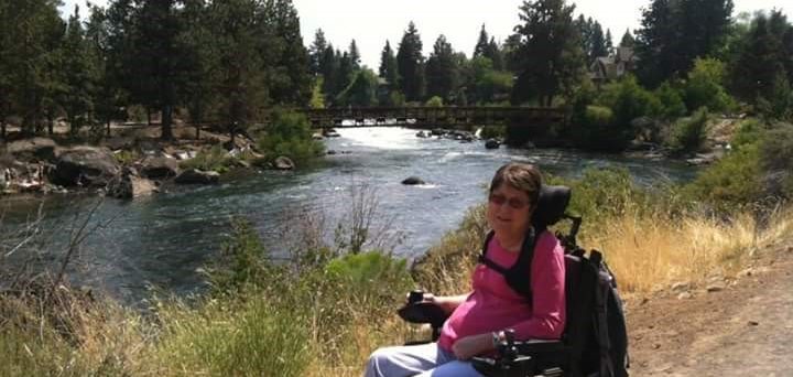 Shelley P. seated in her wheelchair in front of a stream near a forest.